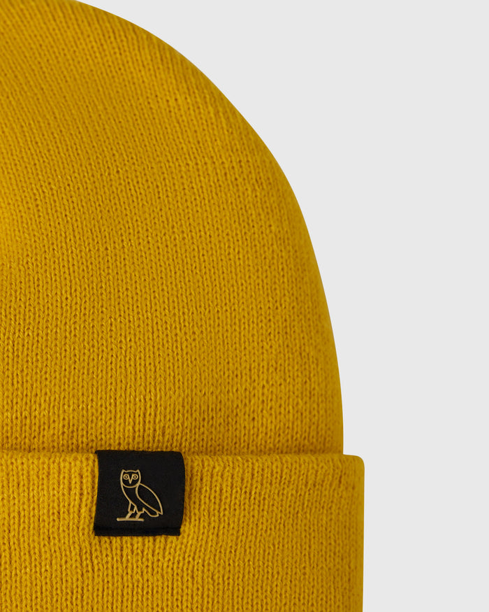 Gold ribbed knit beanie with black woven label featuring owl emblem at cuff.