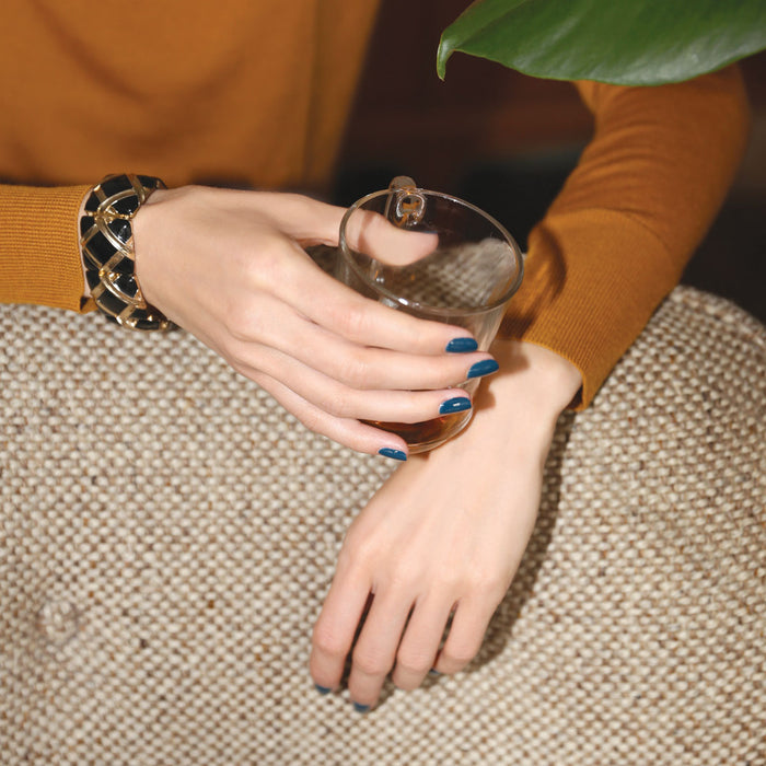 Wearing Manucurist's Dark Clover polish, a person with a black and gold bracelet holds a glass cup on a textured beige surface. They wear a mustard long-sleeve top and part of a green leaf is visible above their hands.