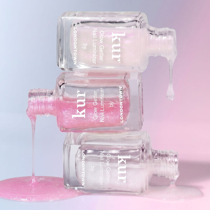 Three clear bottles of LONDONTOWN Glow Getter Nail Luminizer - Diamanté lie stacked, with pink and white polish dripping onto a pastel pink and purple surface, highlighting powerful nail care and an ethereal glow.