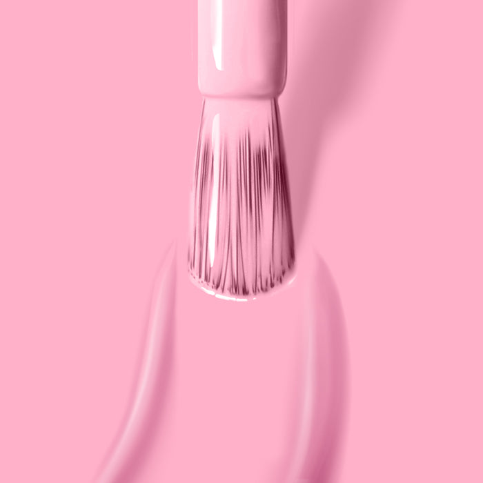 A brush applies LONDONTOWN's Lemonade Pop, a cruelty-free, light pink nail polish, on a matching muted pink background, with smooth strokes and even coverage.