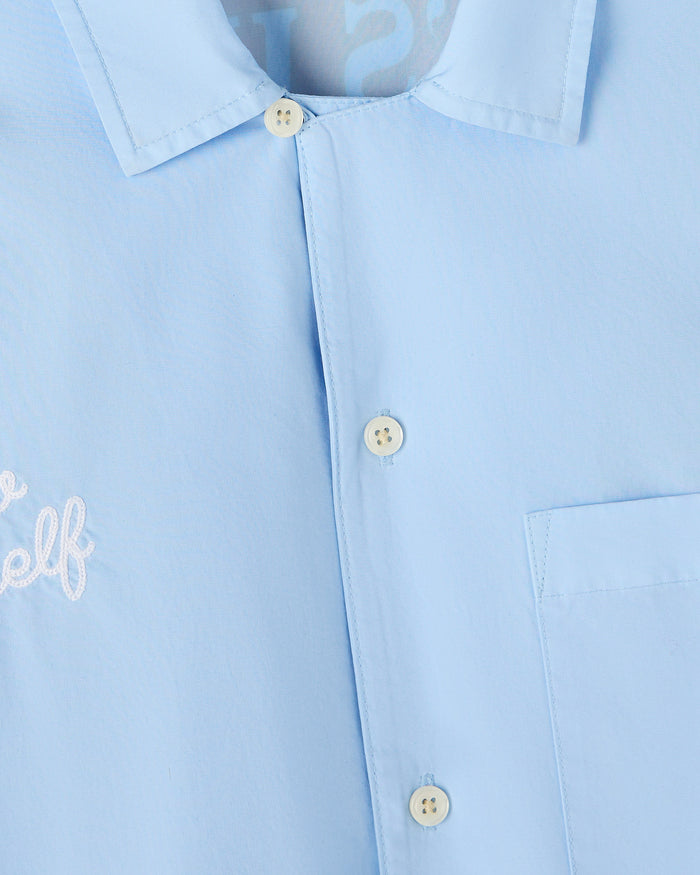 Light blue camp shirt with white pom pom text on chest, spread collar, cream buttons down front, and chest pocket.