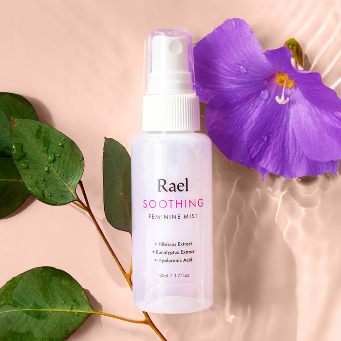 Rael Soothing Feminine Mist with a white spray nozzle on a water background with purple flowers and green leaves.
