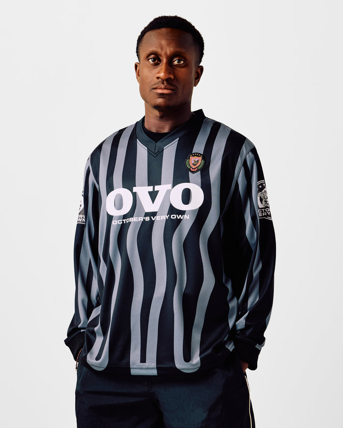Black and gray vertical striped long-sleeve soccer jersey with white sponsor text and chest crest.