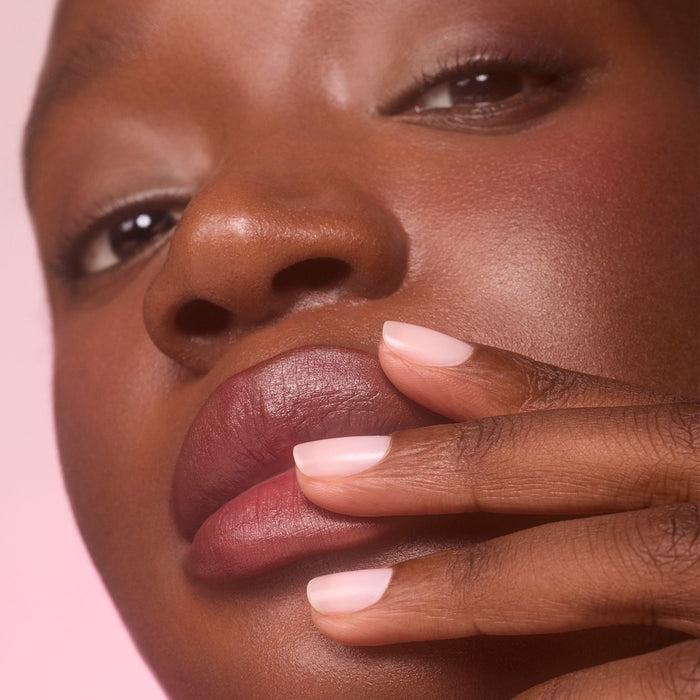 A close-up of a person with dark skin touching their lips with manicured fingers. Their short nails are polished with Manucurist Active™ Blur, a light pink blurring care polish that creates a flawless matte finish. The background is softly blurred in a pink shade.