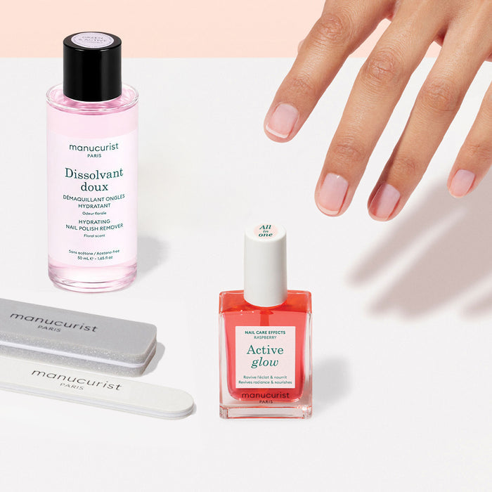 A hand with manicured nails is reaching toward the Manucurist Active Discovery Kit, featuring nail care essentials like nail polish remover, nail polish, and nail files—all arranged on a light surface—perfect for hydrating nails and cuticles as part of your flawless manicure routine.