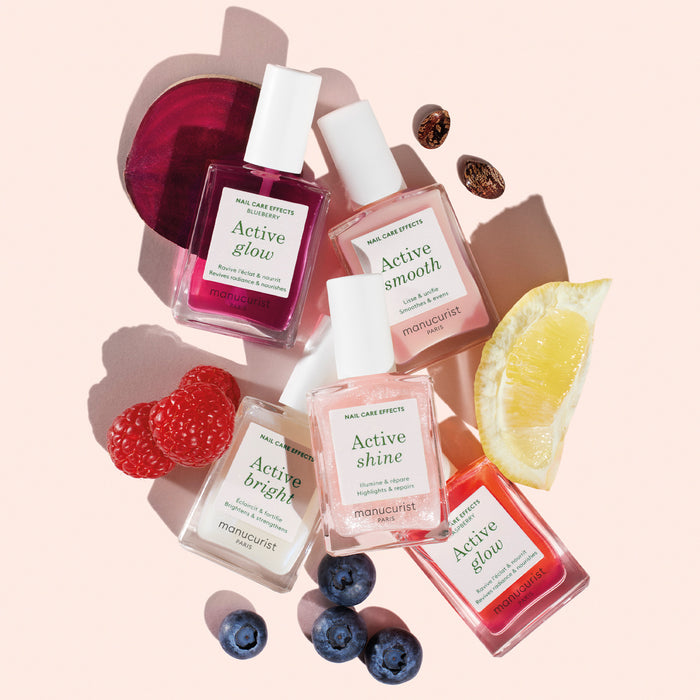Five bottles from the Manucurist Active Discovery Kit are displayed on a pale pink background, surrounded by fresh raspberries, blueberries, lemon, and beetroot.