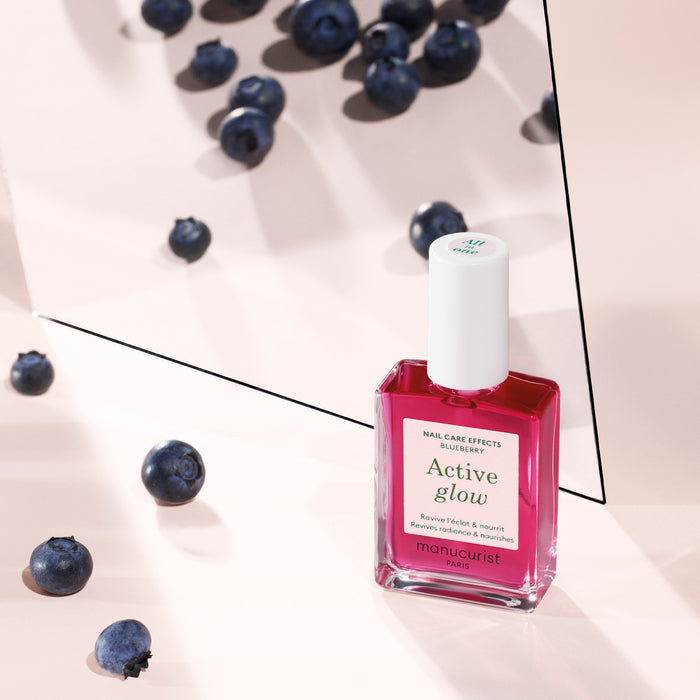 A bottle of Manucurist Active™ Glow Blueberry nail care polish rests on a pink surface surrounded by scattered blueberries, with a reflective mirror displaying the back of the bottle.