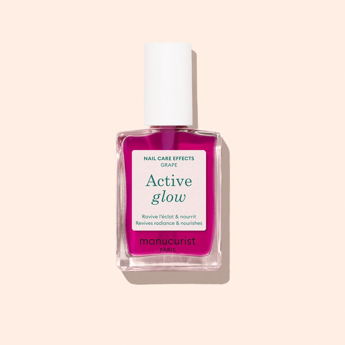 A bottle of Manucurist Active™ Glow Grape nail care polish, featuring a white cap and pink liquid, designed to enhance your natural shine, displayed against a light beige background.