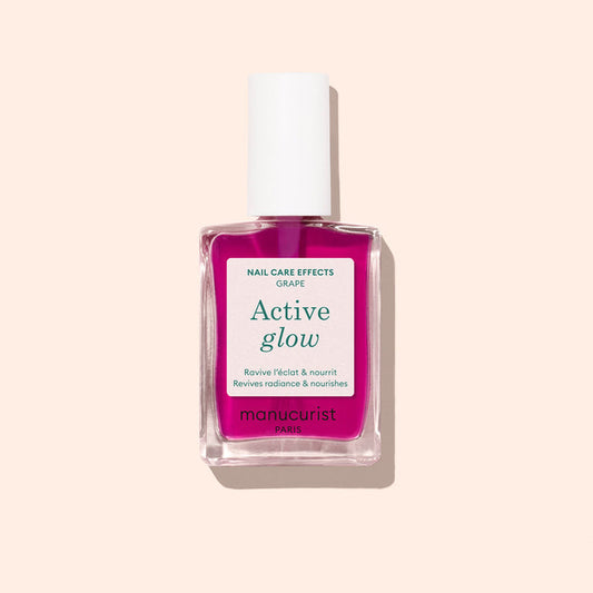 A bottle of Manucurist Active™ Glow Grape nail care polish, featuring a white cap and pink liquid, designed to enhance your natural shine, displayed against a light beige background.