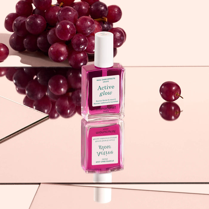 A bottle of Manucurist’s Active™ Glow Grape nail care serum stands on a mirrored surface, with grapes in the background and its reflection visible below, promising natural shine for your nails.