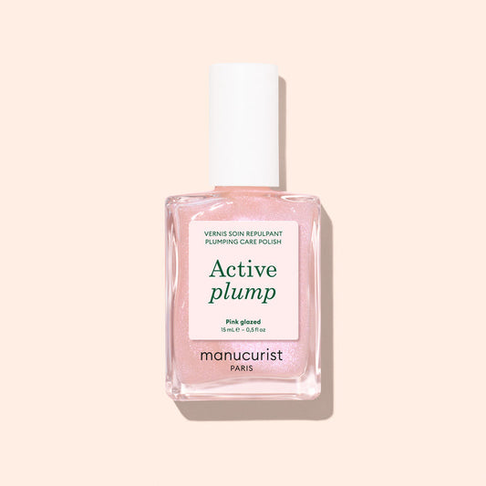 A bottle of Manucurist Active™ Plump Pink Glazed collagen nail polish in a light pink shade, featuring a white cap and displayed against a pale peach background—ideal for hydrated nails and gentle, plant-based nail care.