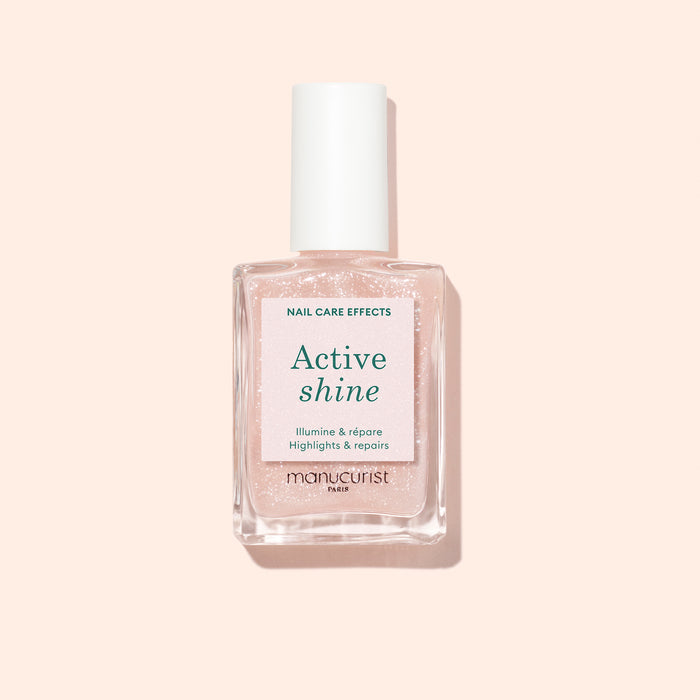 A bottle of Manucurist Active™ Shine plant-based nail polish, featuring a white cap and filled with a light pink, shimmery formula, is displayed on a pale peach background.