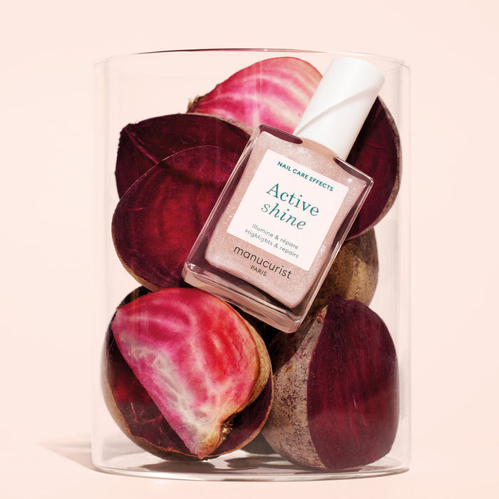 A bottle of Manucurist Active™ Shine—a plant-based nail polish and nail strengthener—rests inside a clear container filled with sliced beets on a light background.