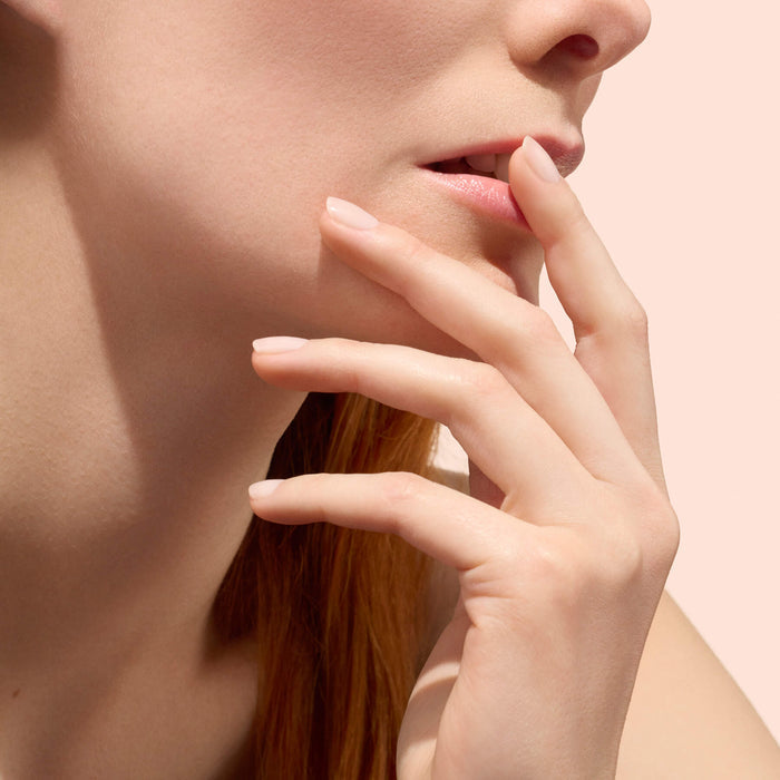 A close-up of a woman touching her lips with her fingers, highlighting smooth skin and natural-looking nails enhanced with Manucurist Active™ Smooth 00, set against a soft pink background.
