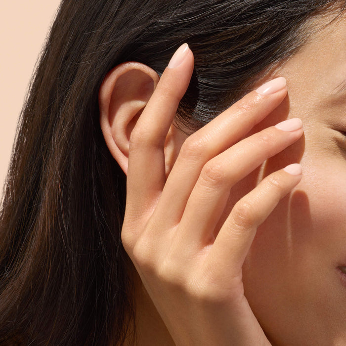 A close-up of a woman’s hand with natural, nude-colored nails—enhanced by Manucurist Active™ Smooth 01 for exceptional nail care—gently touching the side of her face near her ear, set against a soft beige background.
