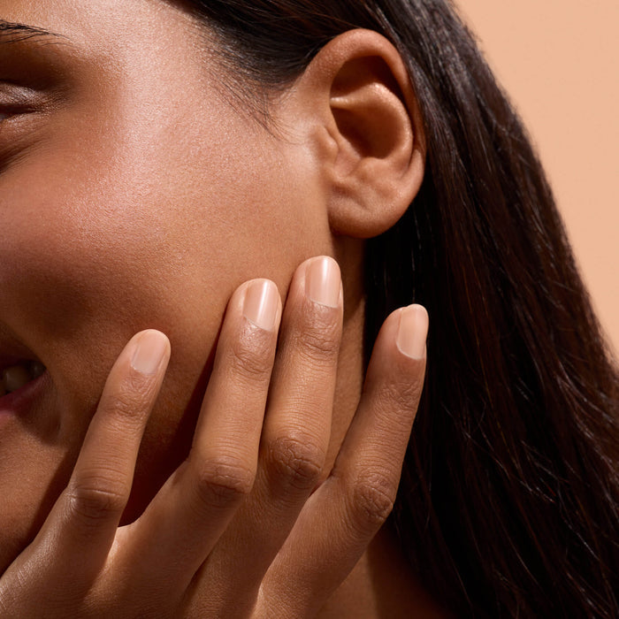 A close-up of a person’s face and hand, showing smooth skin and well-groomed nails—thanks to dedicated nail care with Manucurist Active™ Smooth 02. The person gently touches their cheek and smiles slightly. The background is a soft beige.