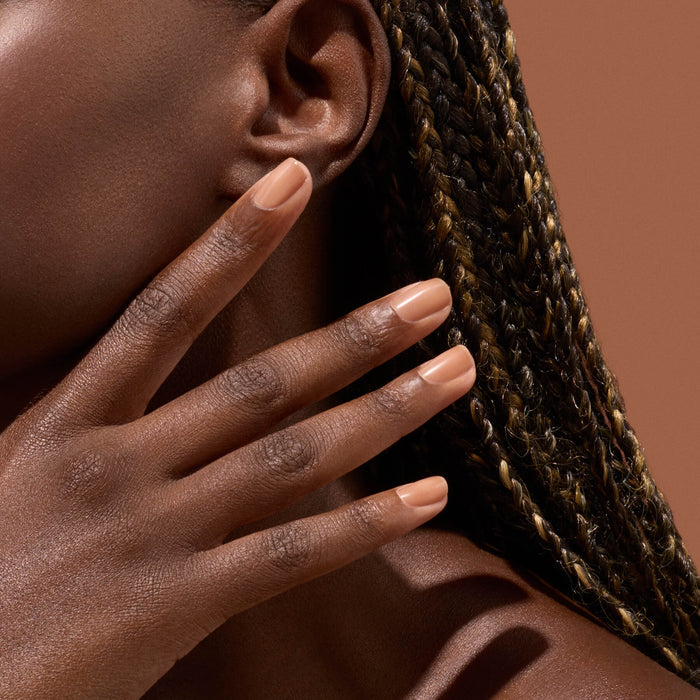 A close-up of a person with braided hair touches their face, displaying neatly manicured nails painted in Manucurist’s Active Smooth 03 nude nail polish against brown skin and a warm brown background.