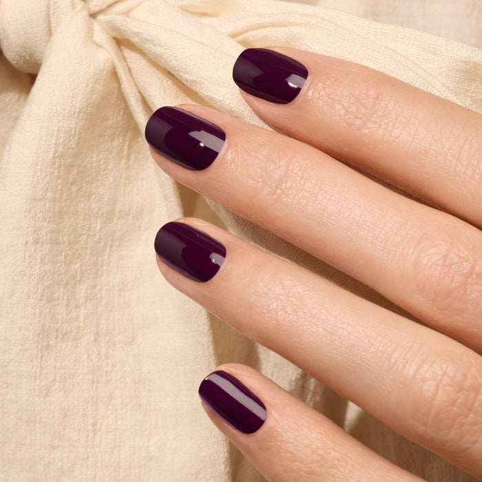 A close-up of a hand with neatly manicured nails painted in Manucurist’s Aubergine gel polish, resting on cream-colored textured fabric tied with a bow.