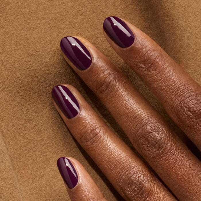 A close-up of a dark-skinned hand resting on brown fabric, featuring neatly manicured nails painted with Manucurist's Aubergine glossy dark purple gel polish.