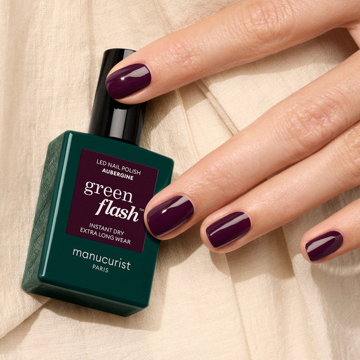 A hand with neatly manicured nails painted in the dark Aubergine shade holds a green bottle of Manucurist's Aubergine polish, resting on cream fabric.