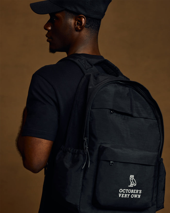 Black backpack with front zippered pocket and white owl logo with "October's Very Own" text on lower back panel.