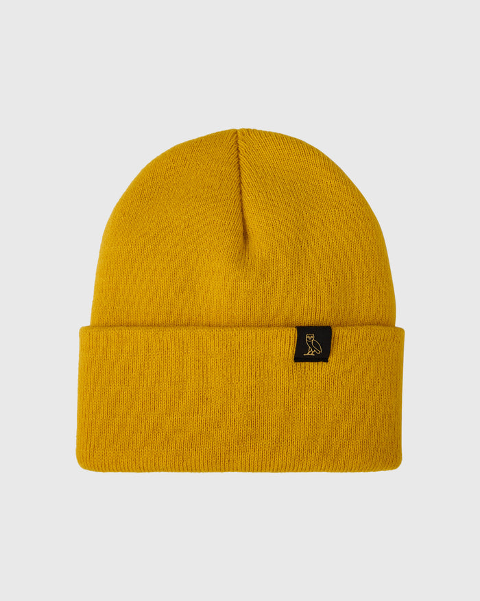 Gold ribbed knit beanie with folded cuff and small black label patch on front.