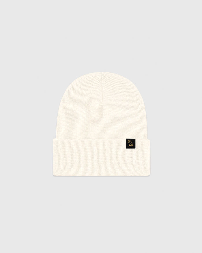 Cream ribbed beanie with small black logo patch on cuff.