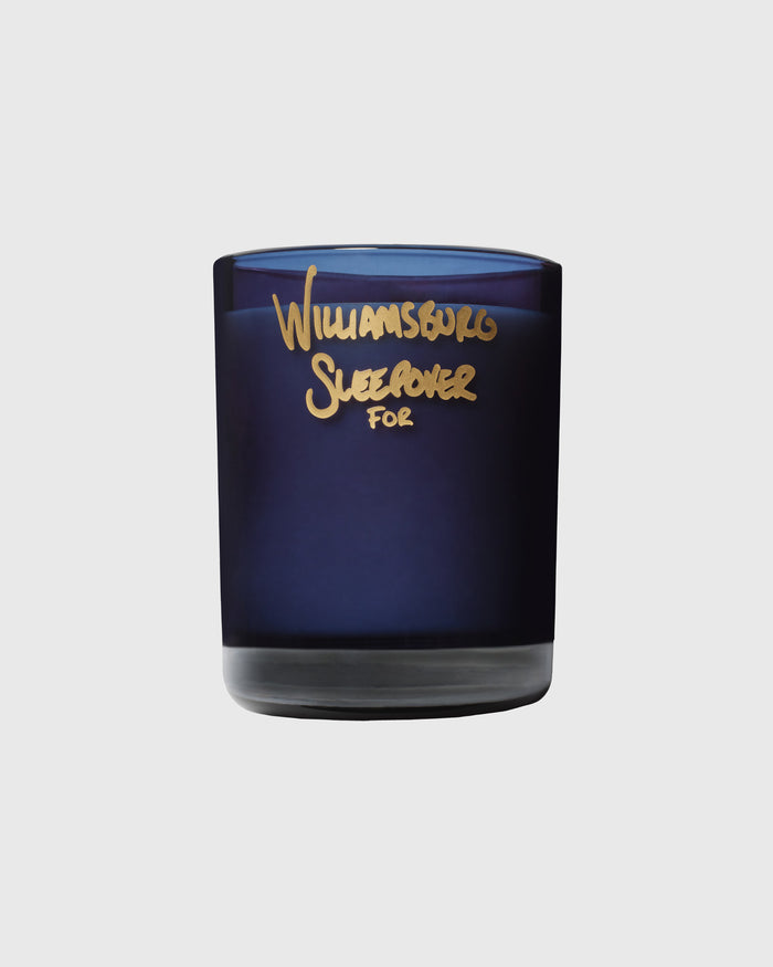 Dark blue glass candle jar with gold foil text reading "Williamsburg Sleepover For" on the label.