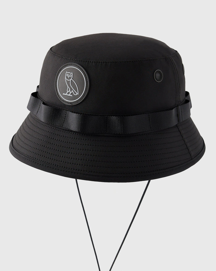 Black boonie hat with owl emblem patch, black webbing band, ventilation eyelets, and chin strap.