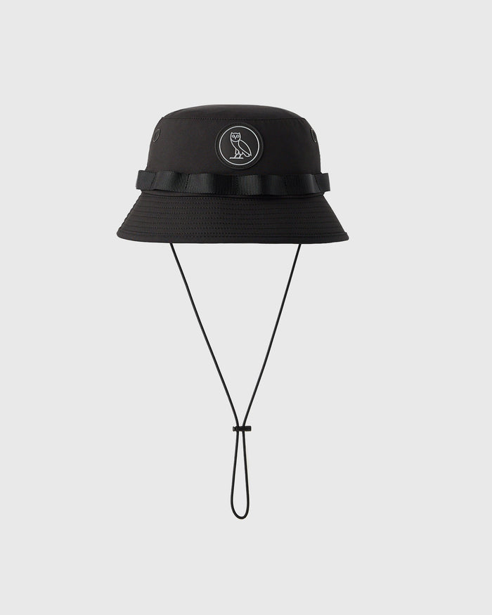 Black boonie hat with wide brim, grosgrain band, circular logo patch, and chin strap.
