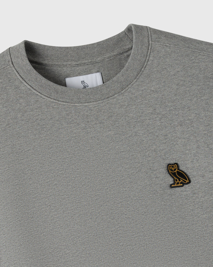 Heather grey crewneck sweatshirt with ribbed neckline and small owl embroidered patch on chest.