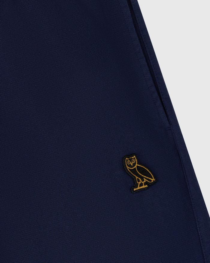 Navy sweatpant with small gold owl embroidery on thigh, ribbed fabric texture visible.