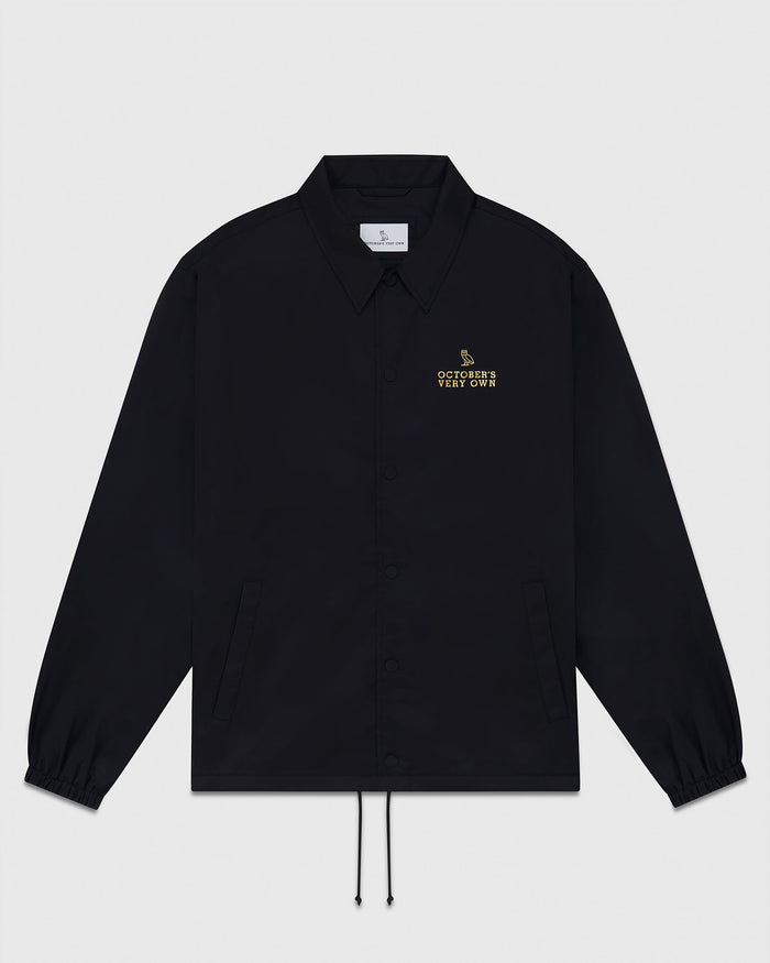 Black coaches jacket with pointed collar, button front, elastic cuffs, and drawstring hem with gold embroidered text on chest