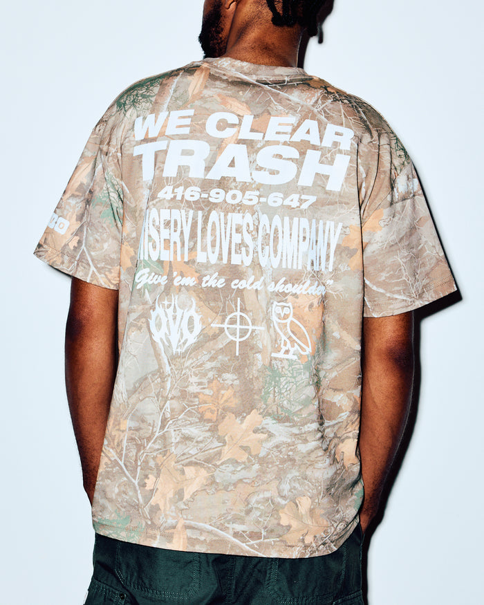 Camouflage tee with white graphic text on back, shown on model from behind wearing dark pants.