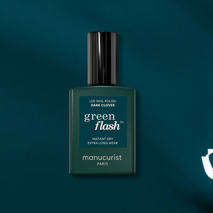 A bottle of Manucurist Dark Clover gel polish is shown against a dark teal background. The dark green bottle, featuring a black cap and white text, highlights this unique blue-green gel nail color.