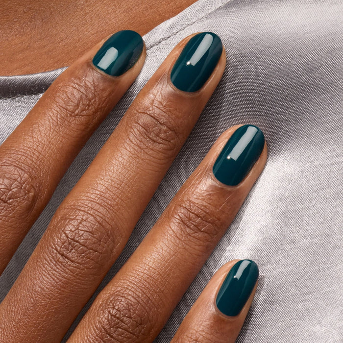 A close-up of a hand with neatly manicured nails painted in Manucurist's glossy blue-green "Dark Clover" polish, resting on light gray satin fabric.