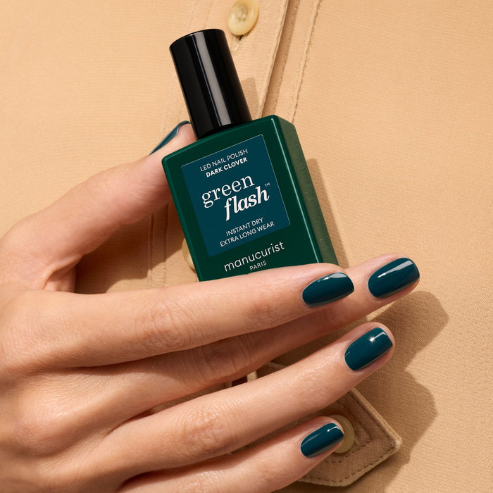 A hand with short, neatly manicured nails painted dark green holds a matching bottle of Dark Clover gel polish from Manucurist against a beige shirt background.
