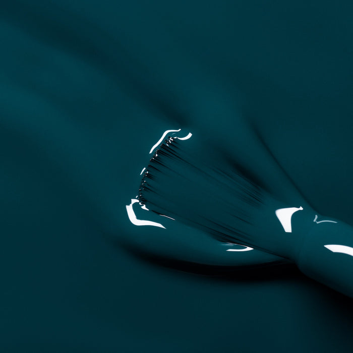 A close-up of a brush applying Manucurist's Dark Clover, a glossy teal gel polish, in smooth strokes with light reflecting off the wet finish.