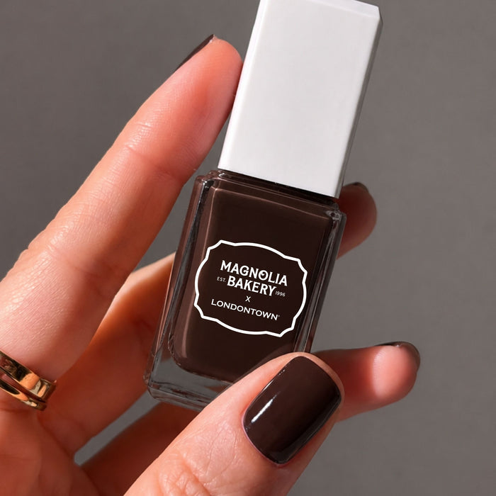 A hand holds a bottle of LONDONTOWN nail polish in the shade “Double Fudge”; the thumbnail is painted with the same rich chocolate brown color.