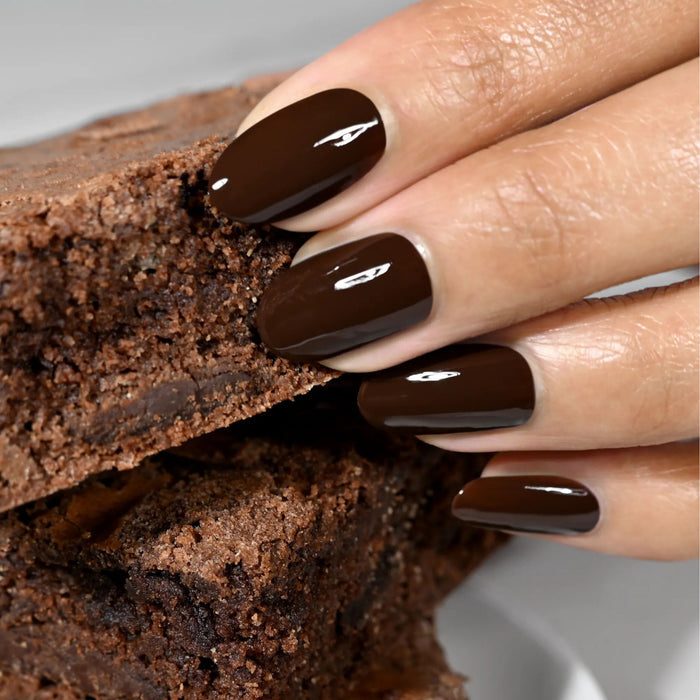A hand with glossy, rich LONDONTOWN Double Fudge chocolate brown nails holds a chocolate brownie against a light background.