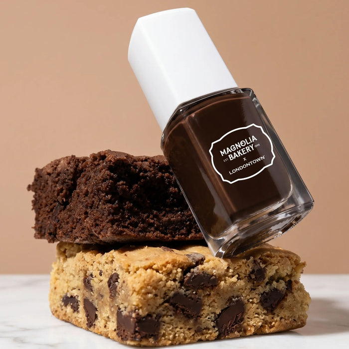 A bottle of LONDONTOWN Double Fudge, a rich chocolate brown nail polish, rests atop stacked chocolate brownie and blondie bars against a neutral background.