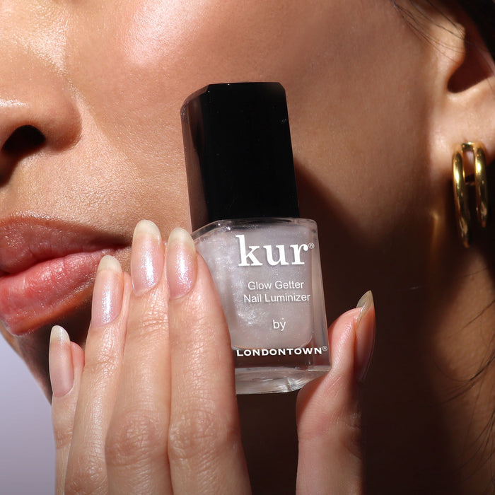 A person with natural nails holds a bottle of LONDONTOWN Glow Getter Nail Luminizer - Diamanté near their face, highlighting its ethereal finish and 360° care for luminous, healthy-looking nails.