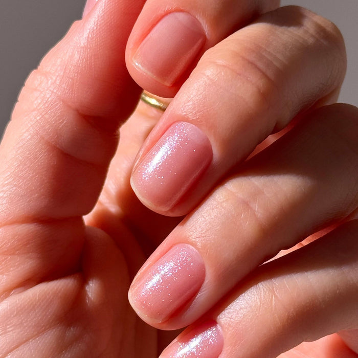 A close-up of a hand with short, natural nails painted in LONDONTOWN's Glow Getter Nail Luminizer - Diamanté, a sheer pale pink polish with an ethereal finish and subtle shimmer.