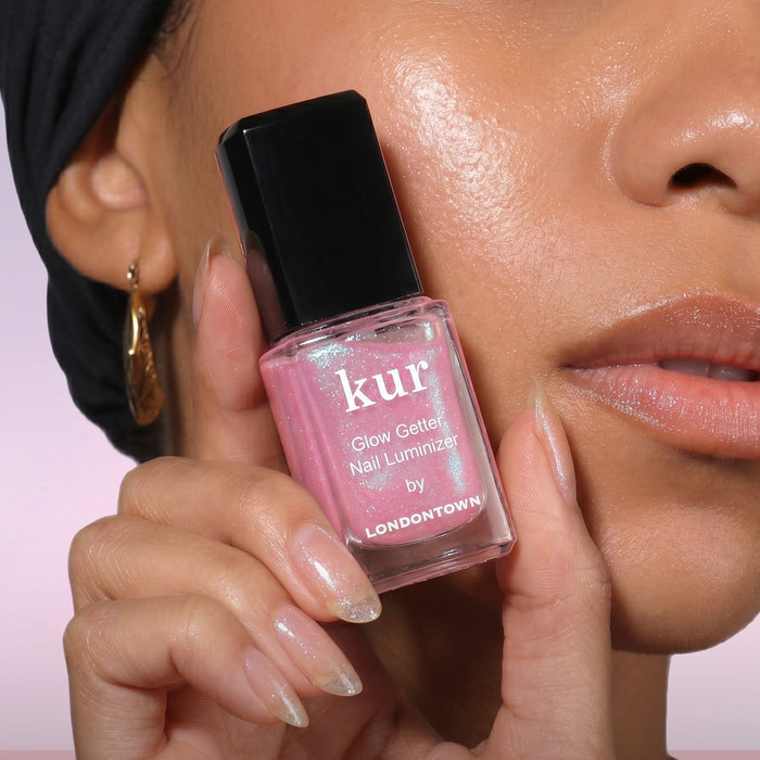 A person holds a bottle of LONDONTOWN Glow Getter Nail Luminizer - Prism near their face, showing off manicured nails and highlighting the product’s radiant finish and powerful nail care benefits.