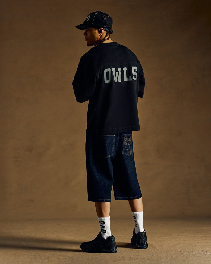 Black heavyweight football tee with "OWLS" text on back, shown with denim shorts and baseball cap.