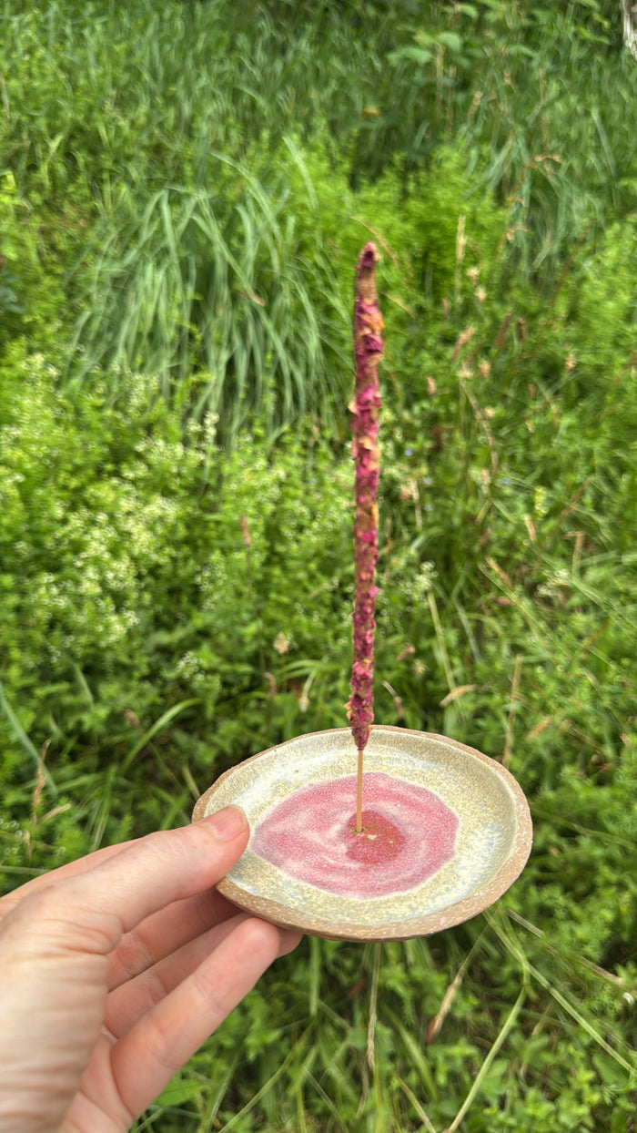 Hand holding a small ceramic dish with a pink substance and a stick over it against a green grass background
