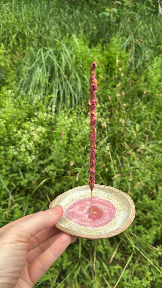 Hand holding a small ceramic dish with a pink substance and a stick over it against a green grass background
