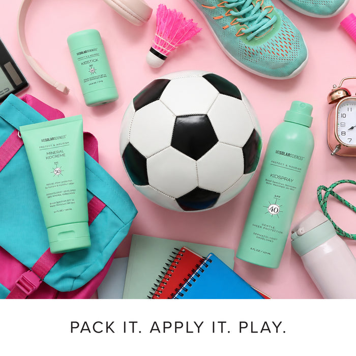 A styled flat-lay of MDSolarSciences kids suncare products in mint green packaging, arranged with a soccer ball, pink backpack, notebooks, sneakers, and other colorful accessories on a pink background. Captioned, "Pack it. Apply it. Play."