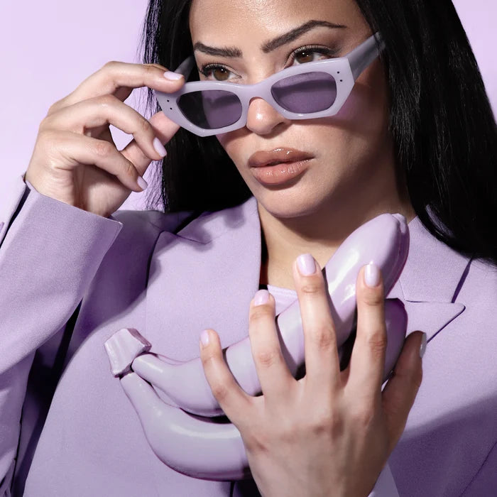 A woman in a lavender blazer and sunglasses holds glossy, lavender bananas against a pastel purple backdrop, capturing the modern style essence of LONDONTOWN's Lavender Illuminating Nail Concealer.