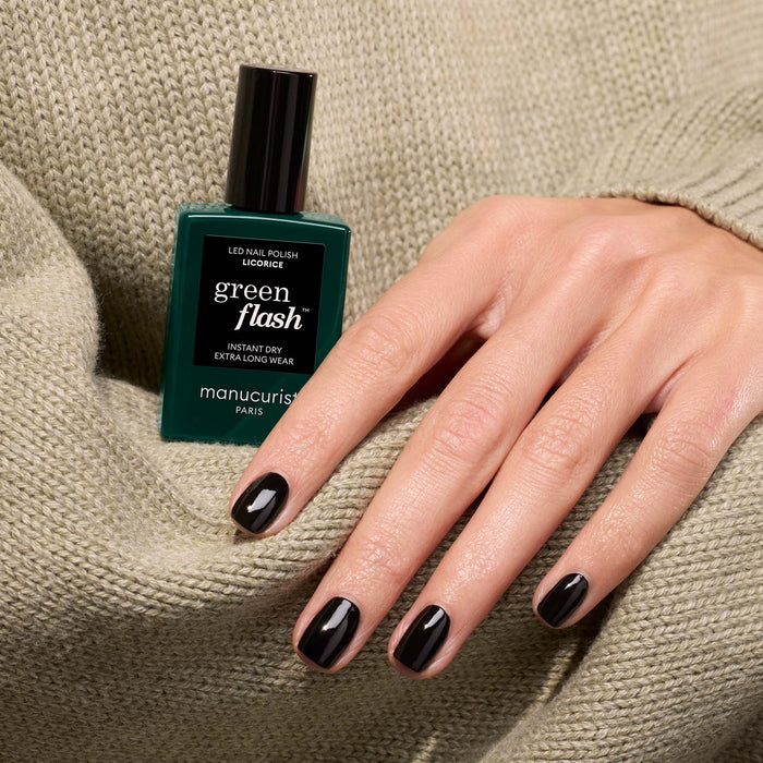 A hand with short nails painted chic black polish rests on a beige knit sweater beside a green Manucurist bottle of Green Flash gel polish in the shade Licorice.